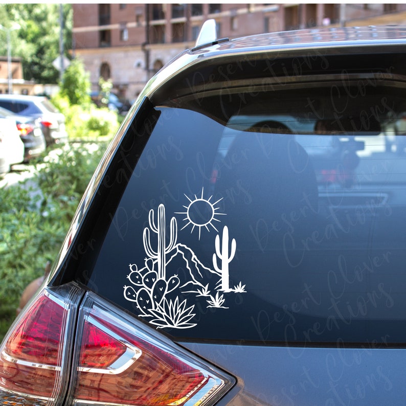 May include: White vinyl decal of a desert scene with cacti, a mountain range, and a sun. The decal is on the back window of a silver car.