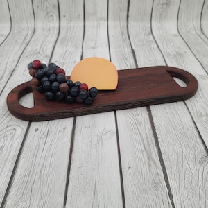 May include: A long, dark brown wooden serving board with two handles. The board has a half-circle of orange cheese and a cluster of dark purple grapes on it.