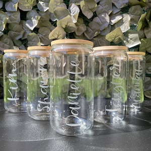 Może przedstawiać: Pięć przezroczystych szklanych słoików z bambusowymi pokrywkami, każdy z innym imieniem wygrawerowanym z przodu: Kim, Shawn, Danielle, Taylor i Naomi.