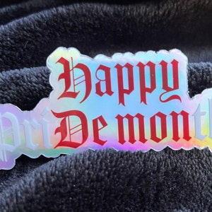 Könnte beinhalten: Ein holografischer Aufkleber mit dem Text "Happy Demon Month" in roten und weißen gotischen Buchstaben.