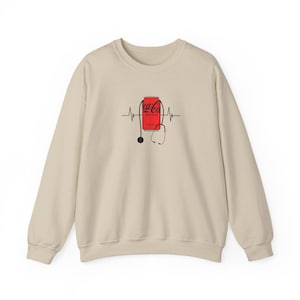 Könnte beinhalten: Beiges Sweatshirt mit roter Coca-Cola-Dosen-Grafik, Stethoskop und Herzschlaglinien-Design. Das langärmlige Oberteil ist aus weichem Material gefertigt.