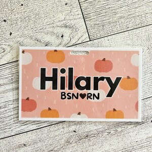 May include: A rectangular name badge with a pink background and a pattern of pumpkins in orange and white. The name "Hilary" is printed in large black letters, with "BSN RN" below.