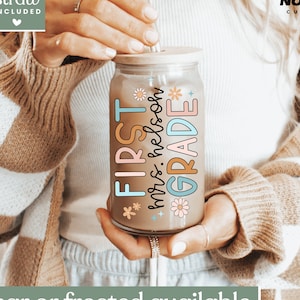 May include: A clear glass tumbler with a wooden lid and a straw. The tumbler has a colorful design that says "FIRST mrs. nelson GRADE" with flowers and stars. The tumbler is being held by a person's hand.