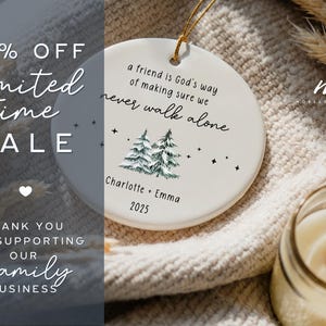 May include: A round white ceramic ornament featuring a gold hanging string. The text reads: a friend is God's way of making sure we never walk alone, Charlotte + Emma, and 2025. It includes a graphic of two green pine trees and small black stars.