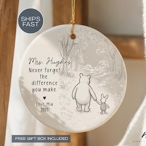 May include: A round, white ceramic ornament with a gold string for hanging. The ornament features a black and white illustration of Winnie the Pooh and Piglet walking. Text reads: "Mrs. Hughes, Never forget the difference you make."