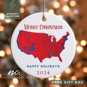 May include: White ceramic Christmas ornament with a red and blue map of the United States. The ornament has the words "Merry Christmas," "Happy Holidays," and "2024" printed on it. A gold string is attached for hanging.