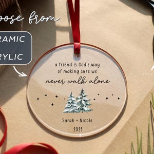 May include: A round, clear acrylic ornament hanging from a red ribbon. The text reads: a friend is God's way of making sure we never walk alone, Sarah + Nicole, and 2025. Below the text are two watercolor-style pine trees and small black stars.