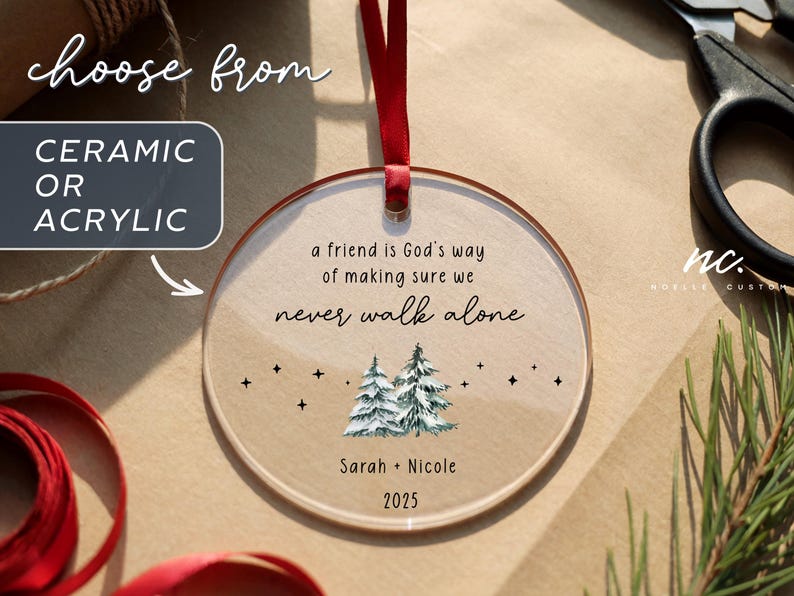 May include: A round, clear acrylic ornament hanging from a red ribbon. The text reads: a friend is God's way of making sure we never walk alone, Sarah + Nicole, and 2025. Below the text are two watercolor-style pine trees and small black stars.
