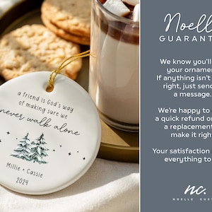 May include: A round white ceramic ornament featuring the text 'a friend is God's way of making sure we never walk alone', a graphic of two pine trees, small stars, the names 'Millie + Cassie', and the year '2024' printed in black ink.