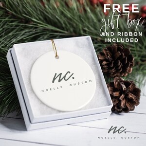 May include: White ceramic Christmas ornament with the text "nc. NOELLE CUSTOM" in gray lettering. The ornament is in a white gift box with a gold ribbon.