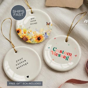 May include: Three white ceramic ornaments with gold string hangers. One features sunflowers and a butterfly, another has the text "Custom Message", and the third says "your custom message" with a heart. The ornaments are round.