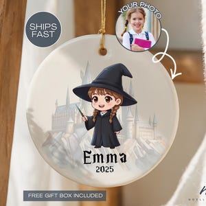 May include: A round ceramic ornament featuring a cartoon illustration of a girl in a witch costume holding a wand. The ornament has the name "Emma" and the year "2025" printed on it. A photo of a girl is in the upper right corner. The background shows a castle.