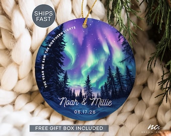 May include: A round ceramic ornament featuring a vibrant illustration of the northern lights above a dark forest silhouette. The text reads THE YEAR WE SAW THE NORTHERN LIGHTS, Noah & Millie, and 06.17.26. It hangs from a gold string.