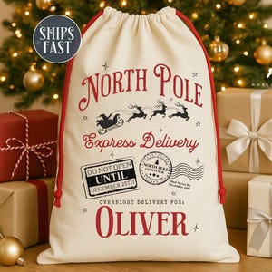 May include: Cream-colored Christmas gift bag with red drawstring and text. The bag reads "North Pole Express Delivery" with a Santa sleigh and reindeer graphic. The bag also has the text "Do Not Open Until December 25th" and "Overnight Delivery for: OLIVER".