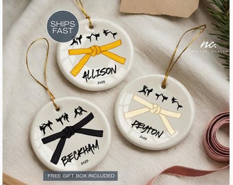 May include: Three white ceramic ornaments with gold hanging strings. Each ornament features a martial arts belt design in yellow, black, and tan, with a name and year. Silhouettes of martial arts figures are above the belt.