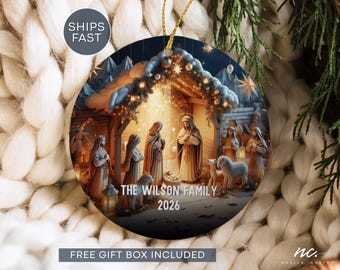 May include: A round ceramic Christmas ornament featuring a detailed nativity scene with figures in a stable, illuminated by warm golden light. The text THE WILSON FAMILY and 2026 is printed in white at the bottom. A gold string is attached for hanging.