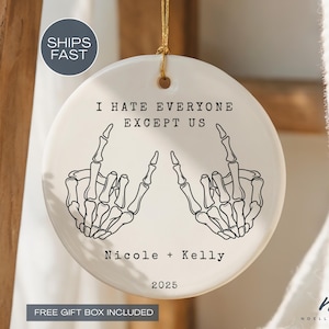 May include: A white ceramic ornament with a gold string for hanging. The ornament features the text "I HATE EVERYONE EXCEPT US" and two skeleton hand illustrations. The names "Nicole + Kelly" and the year "2025" are also printed on the ornament.