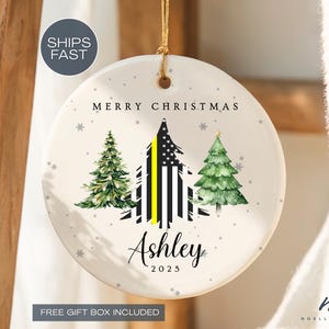 May include: A round, white Christmas ornament with a gold string. It features three green Christmas trees and a stylized tree with black and white stripes and a yellow stripe. The ornament says "Merry Christmas", "Ashley", and "2025".