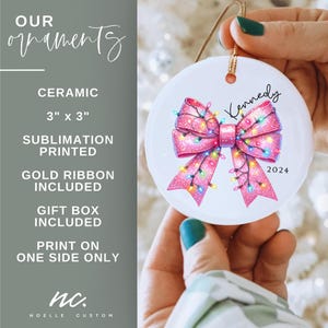 May include: A white ceramic Christmas ornament with a pink bow decorated with colorful lights. The ornament is personalized with the name "Kennedy" and the year "2024".
