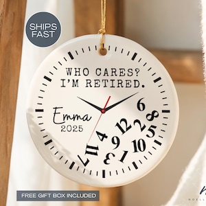 May include: A white ceramic clock-shaped ornament with the text "WHO CARES? I'M RETIRED." and "Emma 2025." The clock face has black numbers and hands, with a red second hand. The ornament is hanging by a gold string.