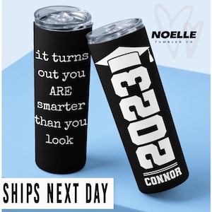 May include: Two black stainless steel tumblers with white text. One tumbler says "it turns out you ARE smarter than you look". The other tumbler says "2023" with a graduation cap above the number and "CONNOR" below the number.