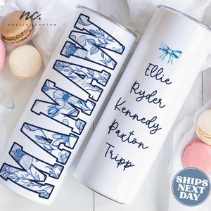 May include: Two white stainless steel tumblers. One features the word "MAMA" in blue floral letters. The other has names in a script font and a blue bow graphic. Macarons in pink, brown, and white are scattered around the tumblers.