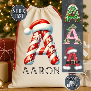 May include: A cream-colored Christmas gift bag with a red drawstring, featuring a large letter "A" designed with red and white candy cane stripes, snowflakes, and a Santa hat. The name "AARON" is printed below the letter.