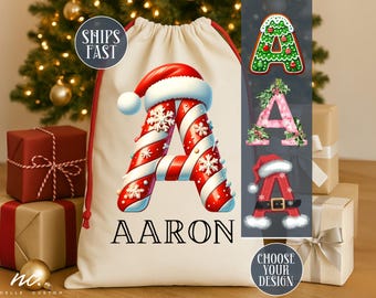 Pode incluir: Uma sacola de presente de Natal creme com um cordão vermelho, apresentando uma grande letra "A" projetada com listras vermelhas e brancas de bengala doce, flocos de neve e um chapéu de Papai Noel. O nome "AARON" é impresso abaixo.