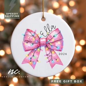 May include: A white ceramic Christmas ornament with a pink bow decorated with colorful lights. The bow has a sparkly finish and the ornament says "Ella 2024".