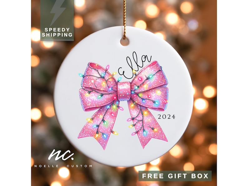 May include: A white ceramic Christmas ornament with a pink bow decorated with colorful lights. The bow has a sparkly finish and the ornament says "Ella 2024".