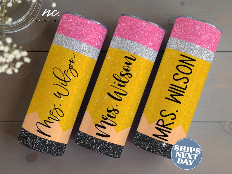 Op de afbeelding: Drie gele en roze glitterbekers met een potloodontwerp en de tekst "Mrs. Wilson" op elk.