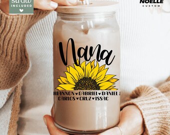 May include: A clear glass tumbler with a wooden lid and straw, featuring the word "Nana" in black script above a yellow sunflower. Below the sunflower are names. The tumbler is held by a person.