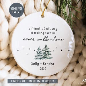 May include: A round white ceramic ornament featuring the black text 'a friend is God's way of making sure we never walk alone', a graphic of two green pine trees, small black stars, and the names 'Sally + Kendra' with the year '2026' below.