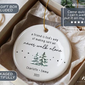 May include: A round white ceramic ornament featuring the black text 'a friend is God's way of making sure we never walk alone', a green pine tree illustration, small black stars, and the names 'Charlotte + Emma' with the year '2025' printed below.