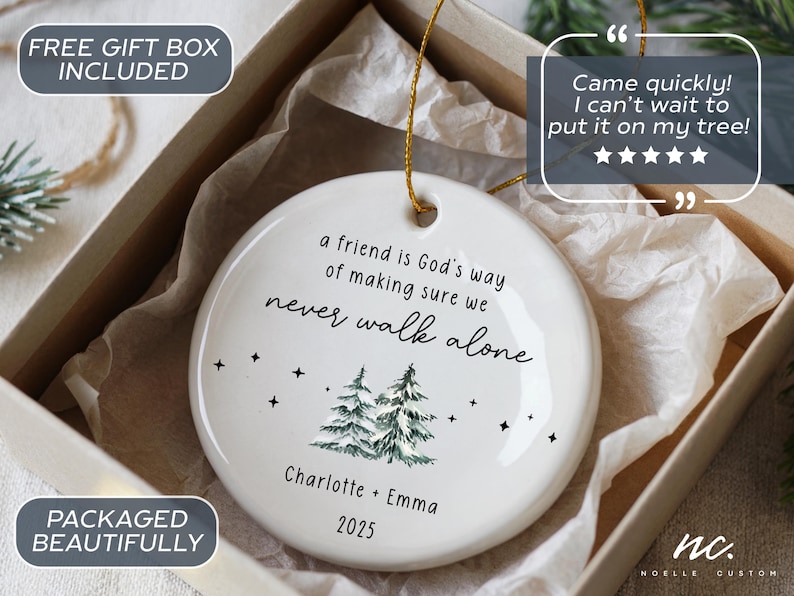 May include: A round white ceramic ornament featuring the black text 'a friend is God's way of making sure we never walk alone', a green pine tree illustration, small black stars, and the names 'Charlotte + Emma' with the year '2025' printed below.