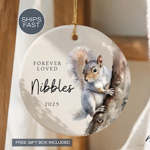 May include: A round ceramic ornament featuring a watercolor illustration of a squirrel on a tree branch. The ornament has the text "Forever Loved Nibbles 2025". A gold string is attached for hanging.