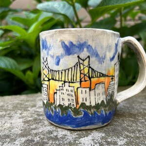 May include: A white ceramic mug with a hand-painted design of a city skyline with a bridge, buildings, and water. The sky is painted in shades of blue and yellow, and the water is blue.