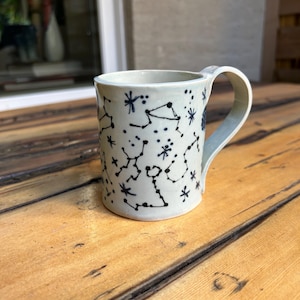 May include: A ceramic mug with a light blue glaze, adorned with a constellation pattern in dark blue. The mug has a curved handle and is set on a wooden surface. The design features stars and connecting lines.