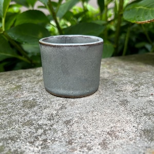 May include: A small, gray ceramic cup with a dark gray rim. The cup is sitting on a gray, textured surface.