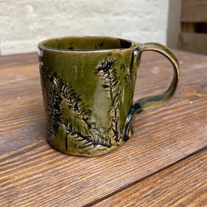 Könnte beinhalten: Eine handgefertigte Keramiktasse mit grüner Glasur und einem strukturierten Farnmuster. Die Tasse hat einen gebogenen Henkel und steht auf einer Holzoberfläche. Die Tasse ist etwa 10 cm hoch.
