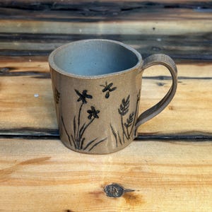 May include: A beige ceramic mug with a black floral design. The mug has a rounded handle and a wide rim.
