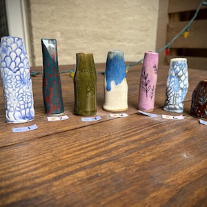 Puede incluir: Una colección de siete jarrones de cerámica en varios colores y diseños. Los jarrones están dispuestos sobre una superficie de madera. Los jarrones incluyen colores azul, verde, rojo y marrón. Algunos jarrones tienen superficies texturizadas y detalles pintados.