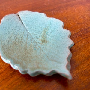 May include: A light blue ceramic leaf dish with a textured surface. The dish is shaped like a leaf with a slightly raised vein running down the center.