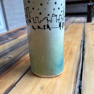 Puede incluir: Un jarrón de cerámica verde con un diseño de ciudad y pájaros. El jarrón tiene un degradado de verde claro a verde oscuro.