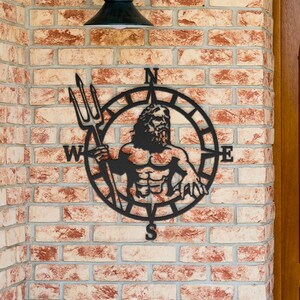 May include: Black metal wall art featuring a compass with a depiction of Poseidon, the Greek god of the sea, holding a trident. The compass points north, south, east, and west.