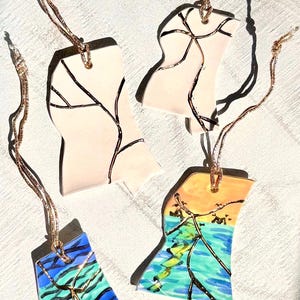 May include: Four decorative ceramic ornaments with gold-colored hanging cords. The ornaments are in the shape of a state, with various designs. Two are light pink with black lines, one is blue and green, and the last one has a sunset and ocean scene.