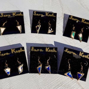 May include: A collection of gold-toned earrings with geometric designs, displayed on black cards. The earrings feature white, blue, and green accents. The cards are labeled with the text "Sara Koch" in gold lettering.