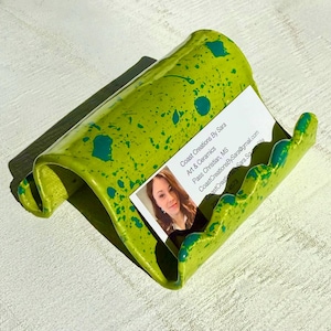 May include: A lime green ceramic business card holder with teal paint splatters. The holder has a curved design and holds a business card with a woman's photo and contact information.