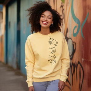 May include: A yellow sweatshirt with a black design of three hands in different positions. The hands are drawn in a simple, minimalist style.