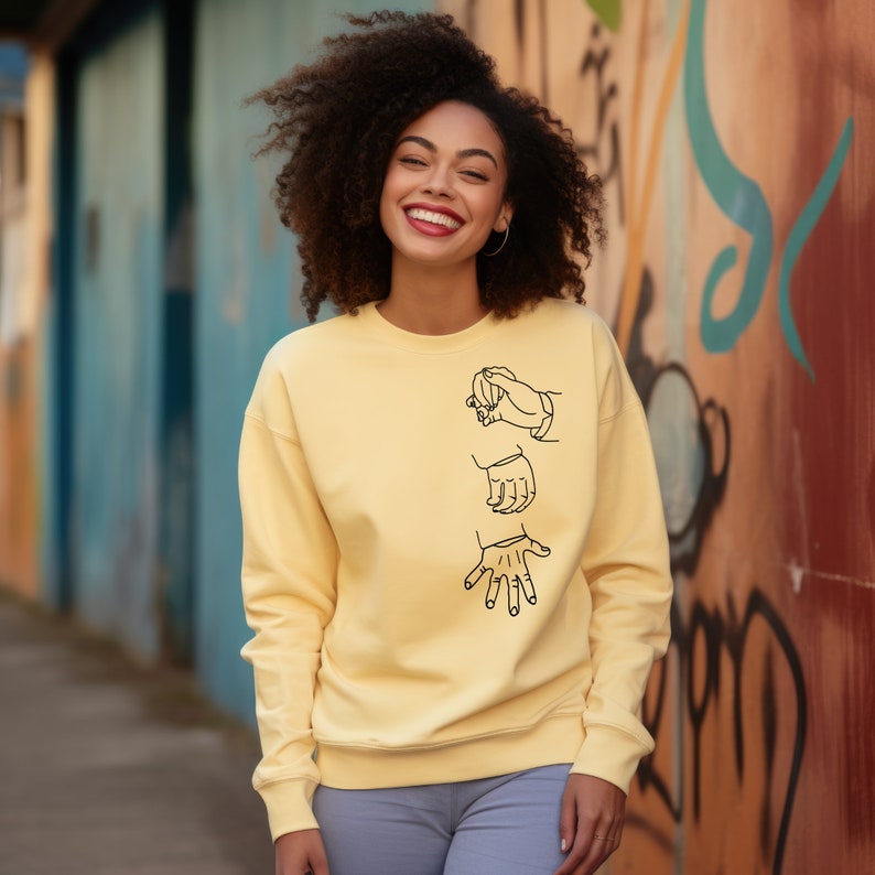 May include: A yellow sweatshirt with a black design of three hands in different positions. The hands are drawn in a simple, minimalist style.